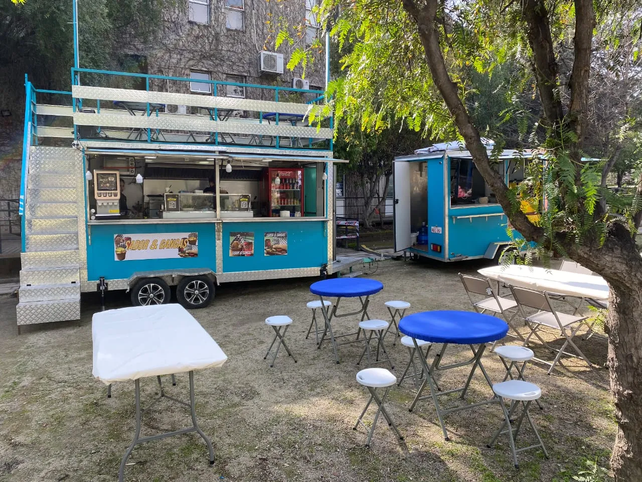 El carro turquesa de Sabores & Gangas con mesas azules bajo los árboles